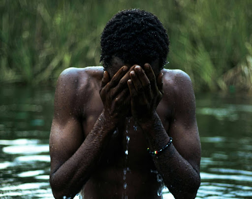 a black man standing in a body of water covering his face with his hands