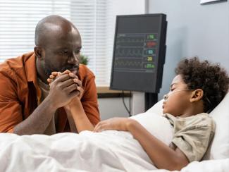 sad father looking at his sleeping son and holding his hand while he is lying in a hospital bed