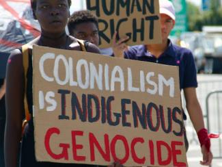 man in black tank top holding sign that says colonialism is indigenous genocide