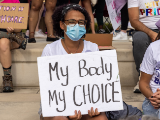 black woman with a face mask on holding a sign that says my body my choice