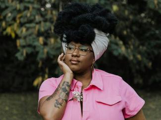 black woman earing a pink shirt and a black and white headwrap with hand on cheek
